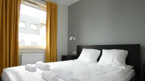 A hotel room with a large bed, two pillows, a nightstand, a lamp, and a window with curtains. - RentPlanet - Apartament Brneńska (Poznań)