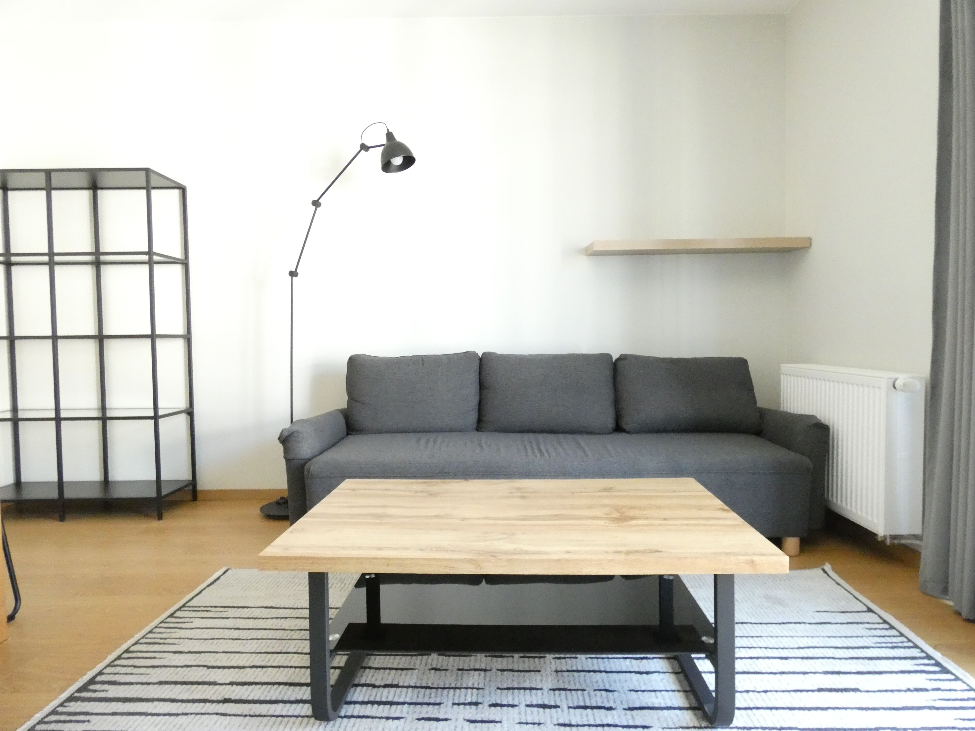 Ett vardagsrum med en grå soffa, ett soffbord i trä, en svart golvlampa och ett vitt element.