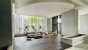 A modern living room with a sofa, chairs, and a small table. There is a window with vertical blinds and a view of greenery outside. - NEW Serenity@Teega ResidenceL2 by UcomeHomestay (Iskandar Puteri)
