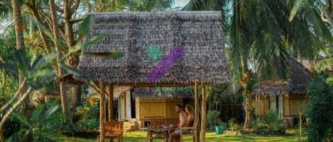 A thatched-roof hut with a wooden bench and table, surrounded by lush greenery and palm trees.