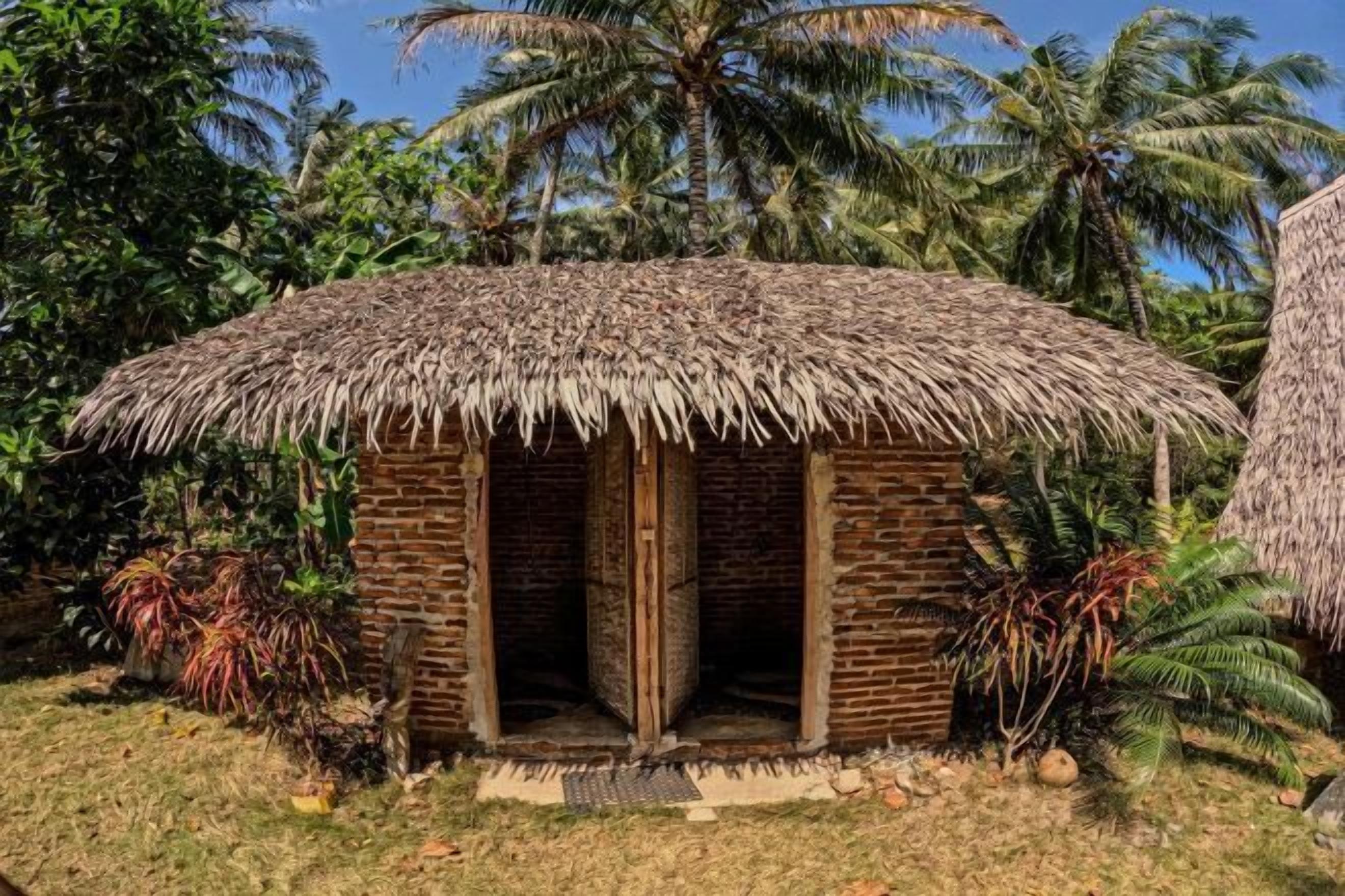 A small, thatched-roof hut with a brick exterior, surrounded by tropical vegetation.