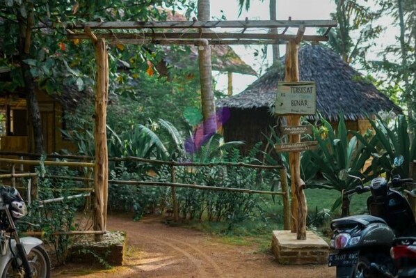 A rustic wooden gate leading to a thatched-roof structure, a signpost with directions, and a motorcycle parked nearby. - Bunga Jabe (Karimunjawa)
