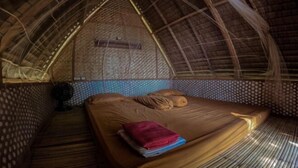A room with a thatched roof, a bed with a blanket and pillows, and a small table with a chair. - Bunga Jabe (Karimunjawa)