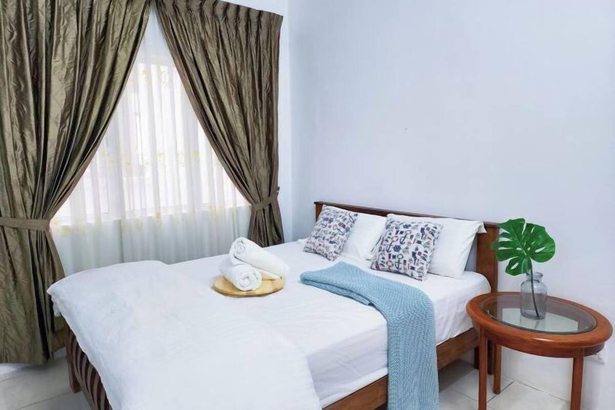 A neatly made bed with a white comforter, pillows, and a blue throw blanket. There is a small round wooden table with a glass top next to the bed, and a potted plant on the table.