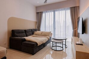 Una habitación de hotel moderna con sofá cama, una mesita redonda, televisor de pantalla plana y un ventanal con cortinas.