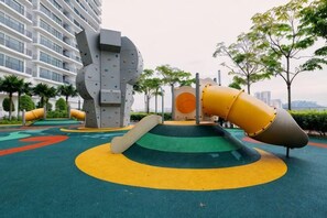 Un parque infantil con toboganes y estructuras de escalada, rodeado de follaje y un edificio al fondo.