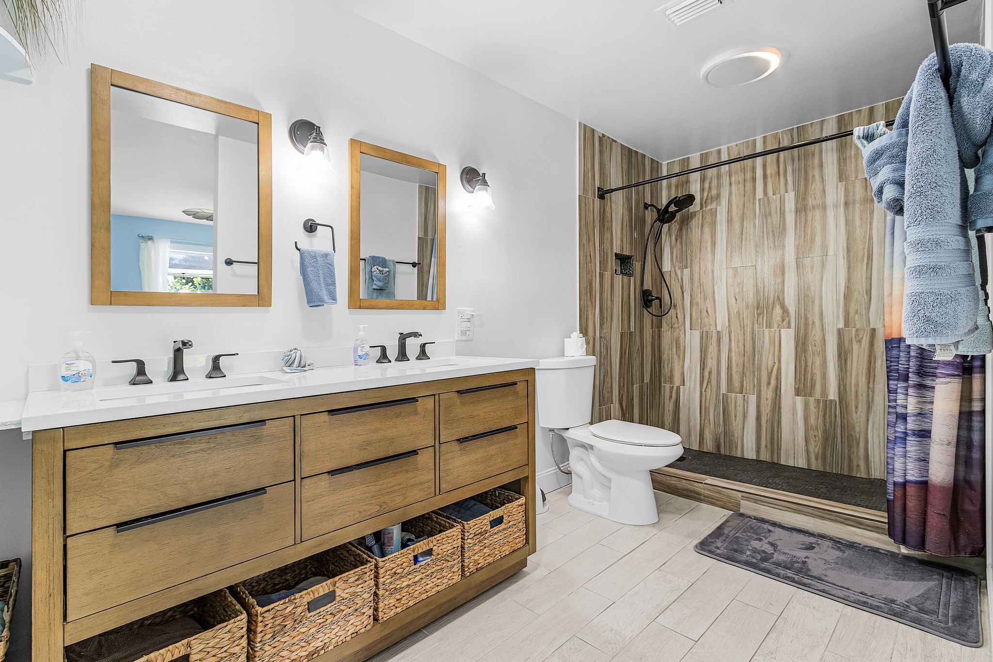 Een moderne badkamer met een houten wastafel, toilet, douche met houten achtergrond en twee wandspiegels.