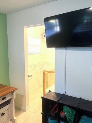 A room with a television mounted on the wall, a wooden table, a white cabinet, and a black storage unit with a blue container.