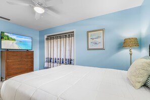 A bedroom with a bed, a television, a framed picture, a lamp, and a window with curtains.