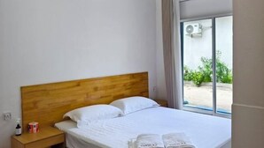 A hotel room with a wooden headboard, a bed with white linens, a nightstand with a bottle and a box, and a view of greenery through a window. - Evelyaa Guest House (Fulidhoo)