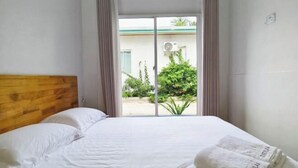 A hotel room with a bed, white linens, a wooden headboard, and a view of greenery and a building outside. - Evelyaa Guest House (Fulidhoo)