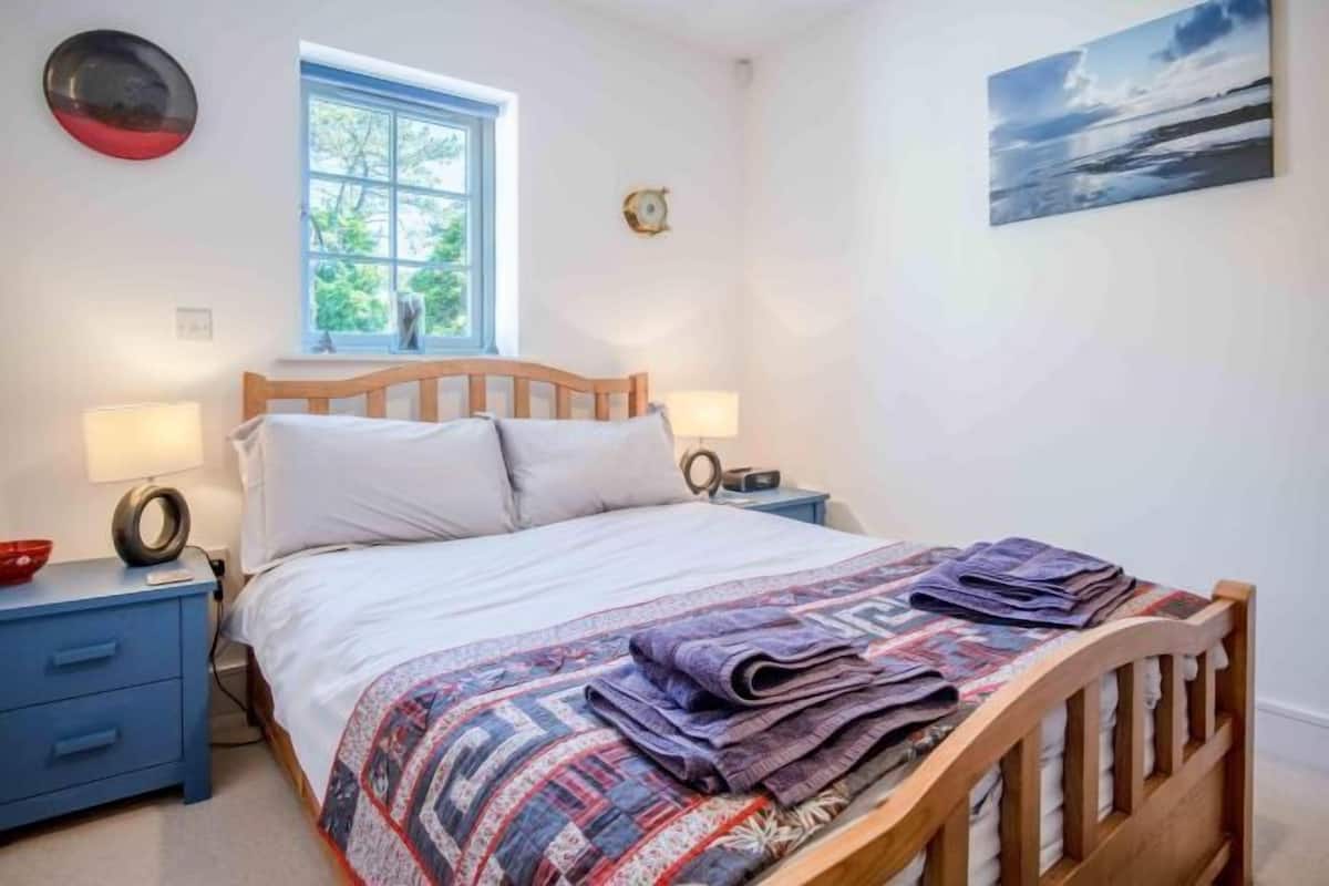 A bedroom with a wooden bed, two bedside tables, a window, a wall clock, and a painting.