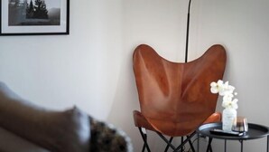 Huoneessa on ruskea nahkainen nojatuoli, pieni pyöreä pöytä, jonka päällä on maljakko ja kaukosäädin, sekä seinällä oleva kehystetty kuva.