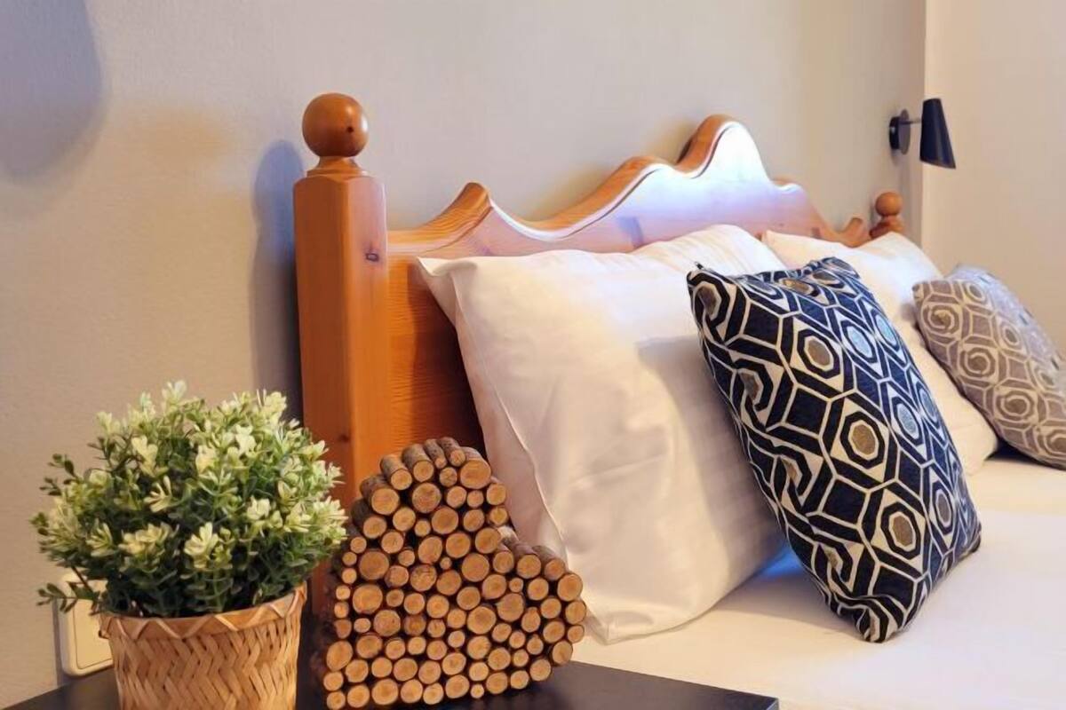 Cama com cabeceira de madeira, almofadas brancas e estampadas, uma planta em vaso e uma pilha de lenha.