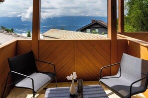 A wooden balcony with two chairs and a table, a vase with flowers, and a view of a house and mountains. - Luxescape-No 2306-Traumaussicht-Ski & Wandergebiet-Paragleiten-Langlauf-Parkplatz (Treffen bei Villach)