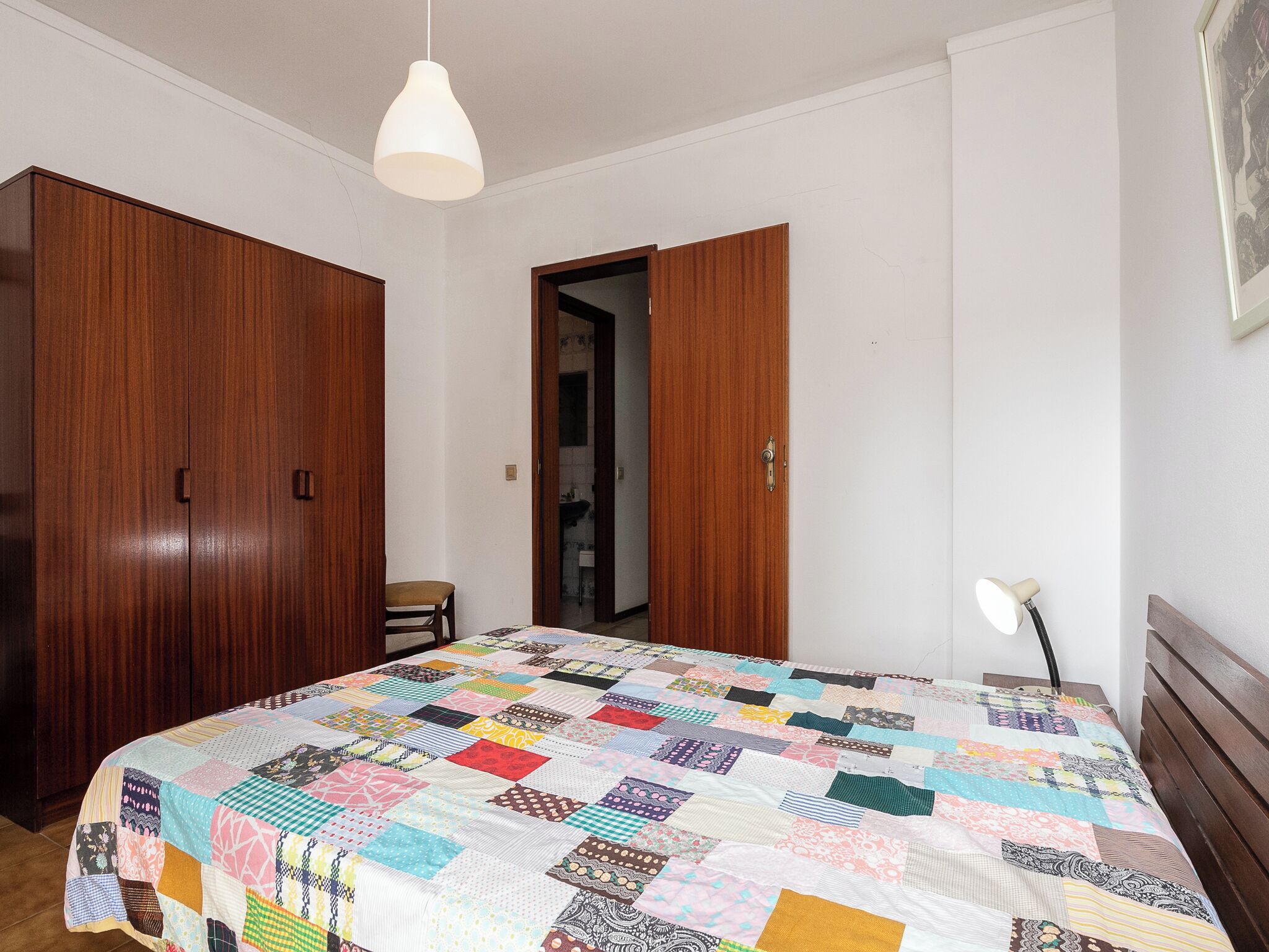 A bedroom with a wooden wardrobe, a bed with a colorful quilt, a white pendant light, and a bedside lamp.