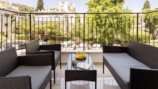 Un balcon agrémenté de meubles en osier, d’une table en verre et offrant une vue sur les immeubles et les arbres.