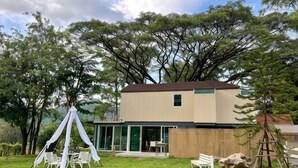 A modern building with a flat roof, large windows, and a covered outdoor seating area with chairs. - Little Tree House(3 bedrooms) (Pak Chong)
