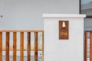 Un portón de madera, una pared blanca con una placa de madera que muestra el número 7718, y una campana montada en la pared.