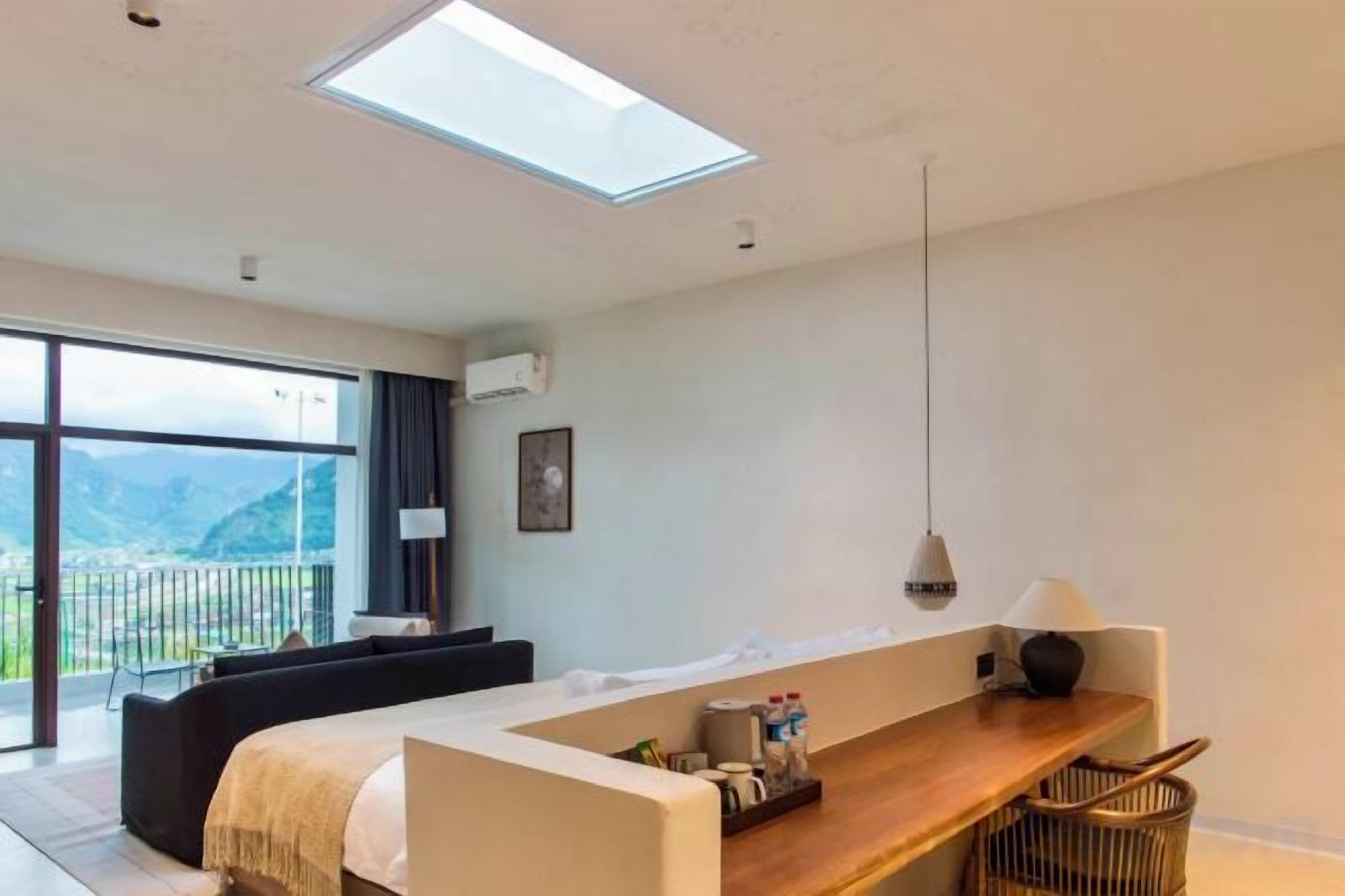 A modern hotel room with a large window, a sofa, a wooden desk, and a view of the outdoors.