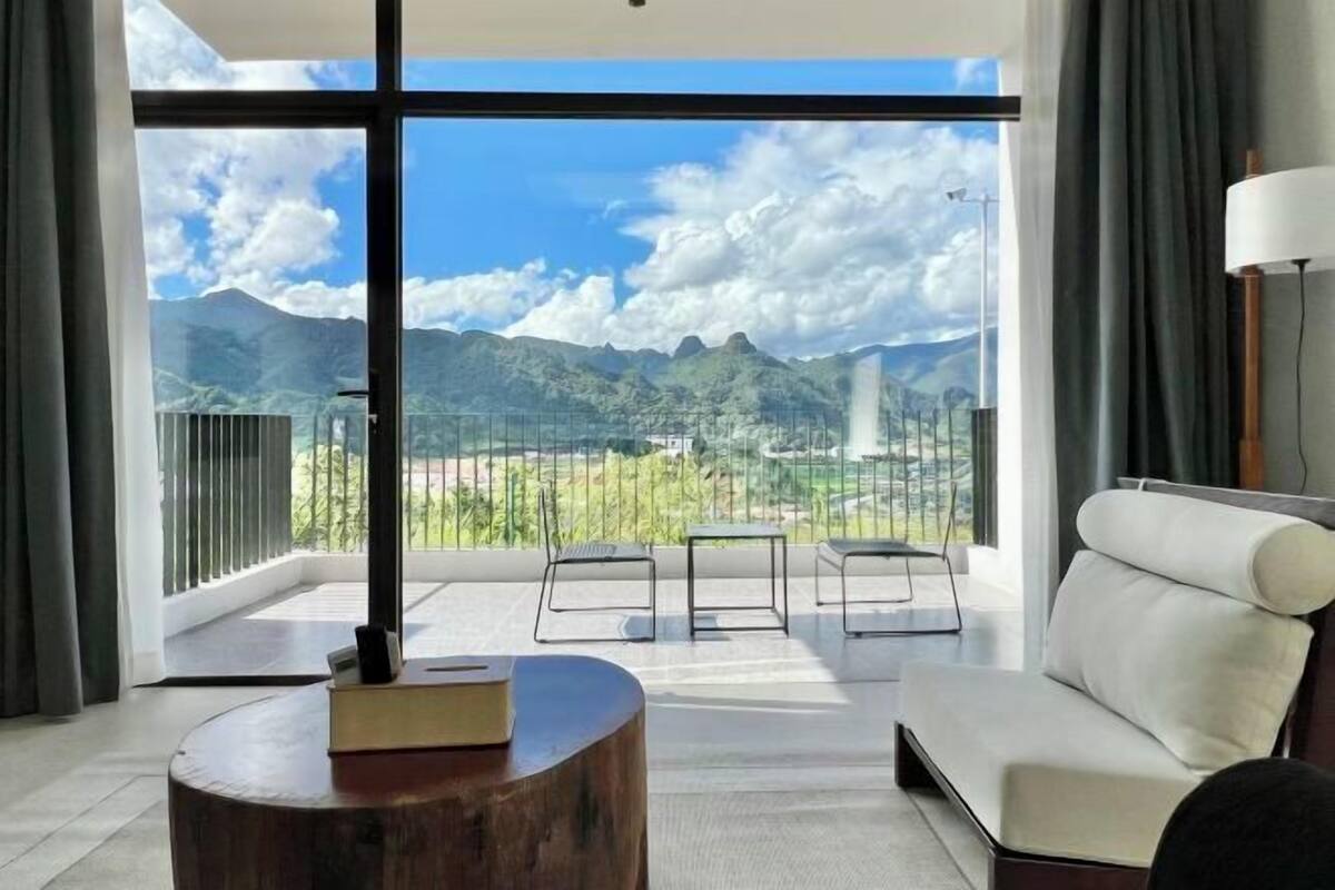 Una habitación moderna con un ventanal grande, un sofá blanco, una mesita pequeña y un taburete de madera.