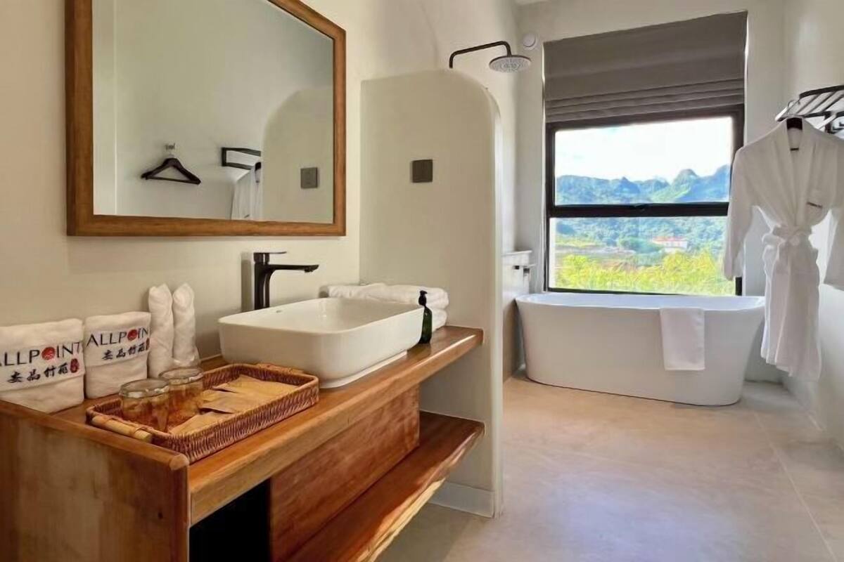 Un baño moderno con un espejo grande, una bañera independiente y una ventana con vistas a zonas verdes.