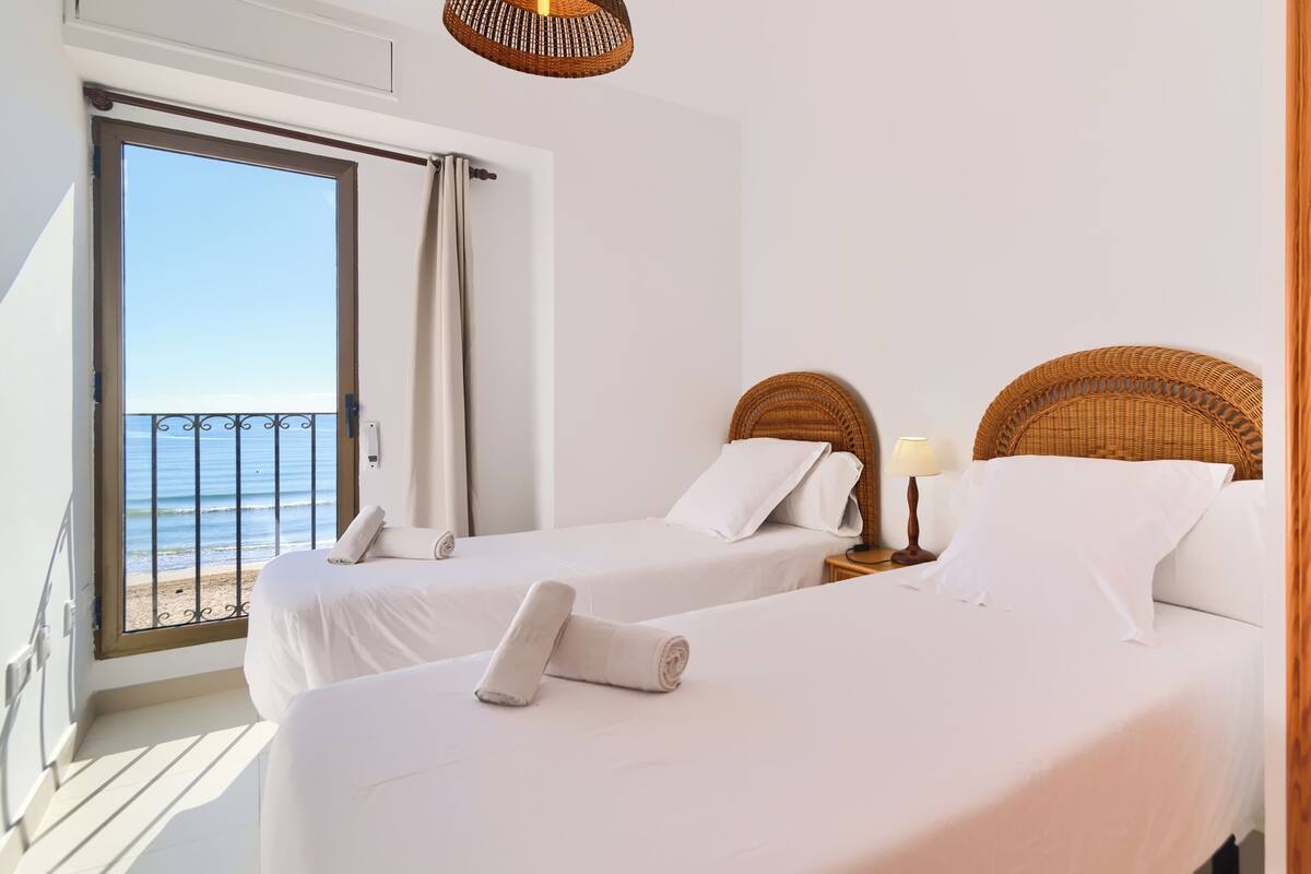 Habitació d'hotel amb dos llits, un balcó i vista al mar, i un capçal de llit de vimet.