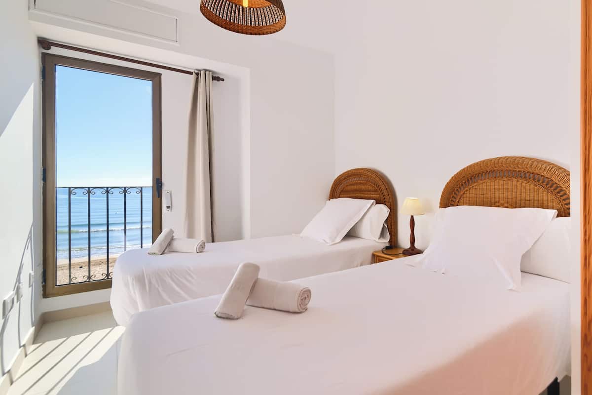 Quarto de hotel com duas camas, varanda e vista para o mar, e cabeceira de vime.