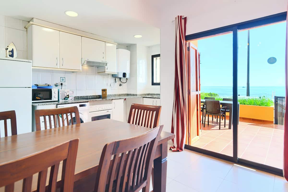 Uma cozinha moderna com armários brancos, uma mesa de jantar com cadeiras e vista para o mar através de uma porta de vidro deslizante.