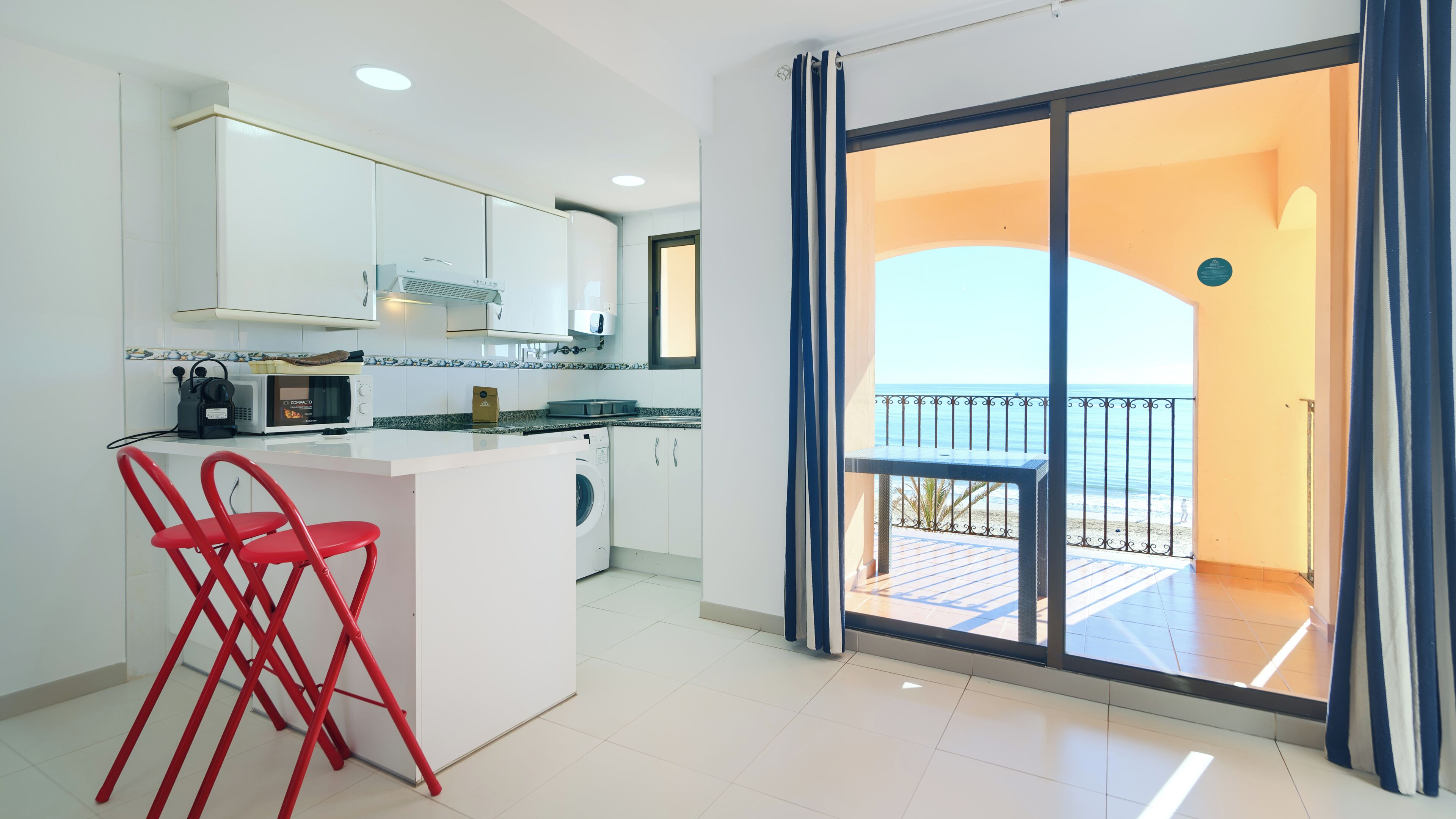 A modern kitchen with white cabinets, a microwave, and a coffee machine. There is a red chair and a small island with a sink. A balcony with a view of the sea is visible through a glass door.