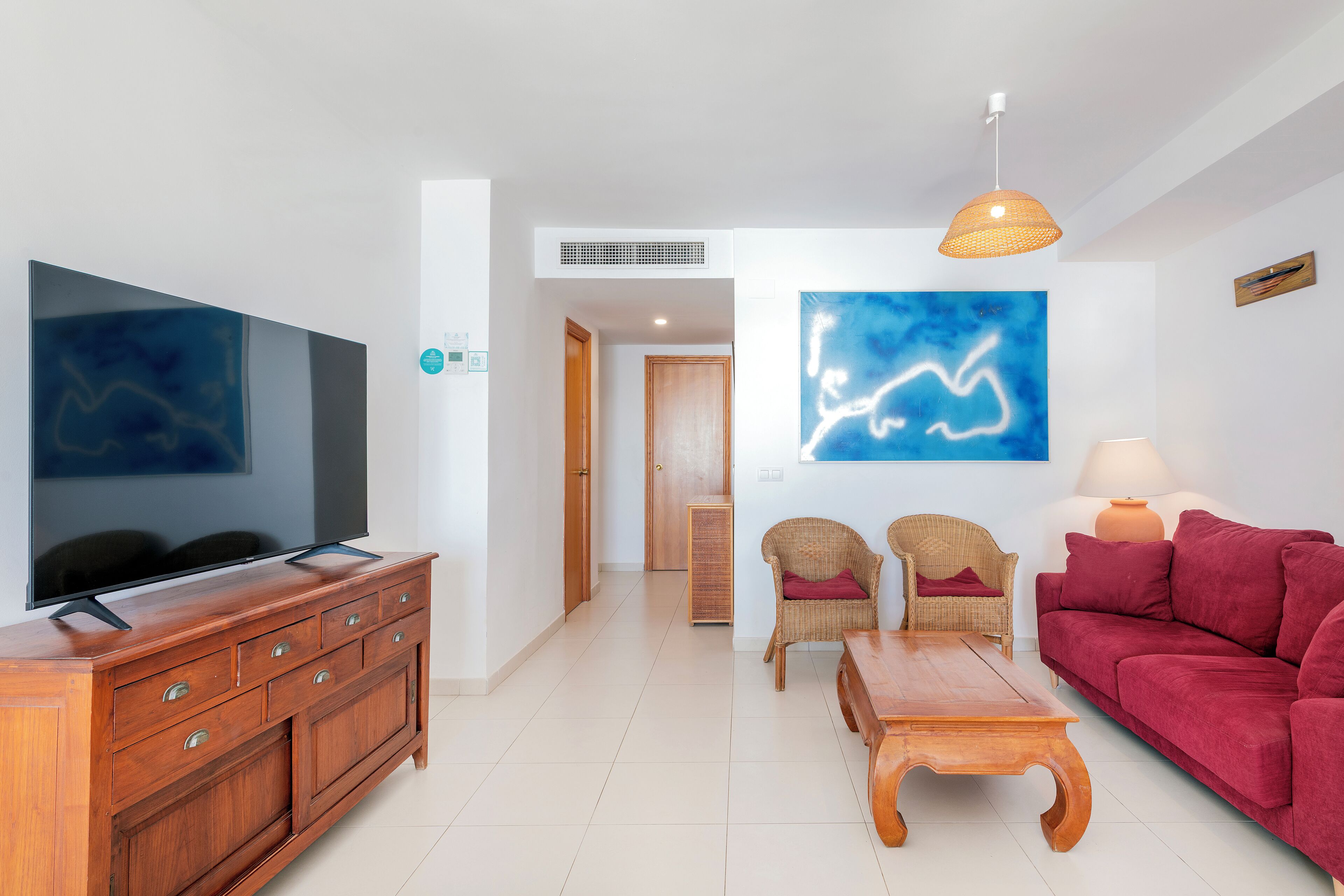 Un salón con una televisión de pantalla plana, un sofá rojo, una mesa de centro de madera, sillas de mimbre y un cuadro abstracto azul.