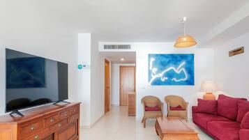 Un salón con una televisión de pantalla plana, un sofá rojo, una mesa de centro de madera, sillas de mimbre y un cuadro abstracto azul.