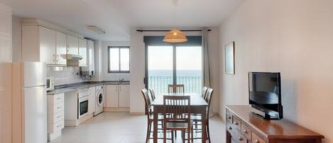Une cuisine moderne avec des meubles blancs, une table à manger avec des chaises, un téléviseur posé sur un meuble en bois et une grande fenêtre offrant une belle vue.