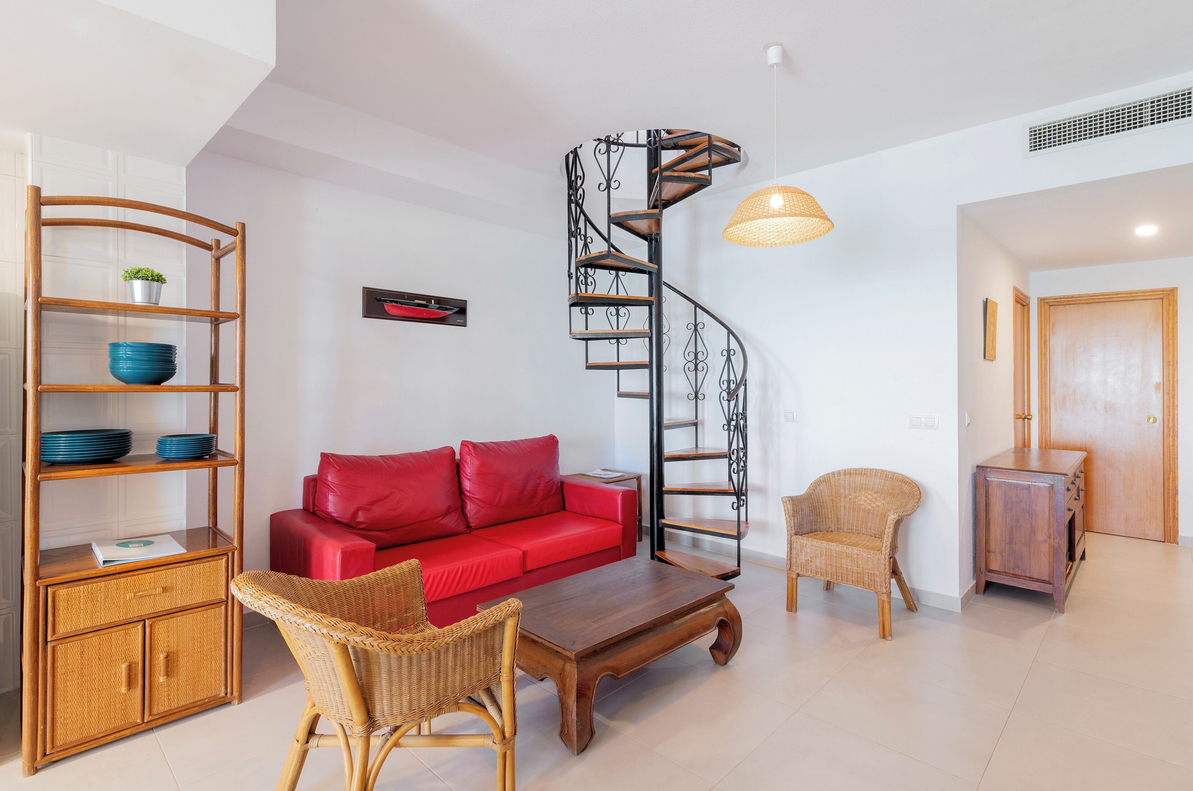 A modern living room with a red sofa, wicker furniture, a wooden coffee table, a spiral staircase, and a hanging light fixture.
