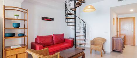 A modern living room with a red sofa, wicker furniture, a wooden coffee table, a spiral staircase, and a hanging light fixture.