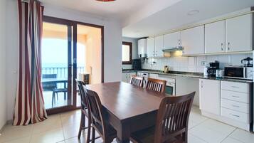 A modern kitchen with white cabinets, a dining table with chairs, and a view of the sea through a sliding glass door.