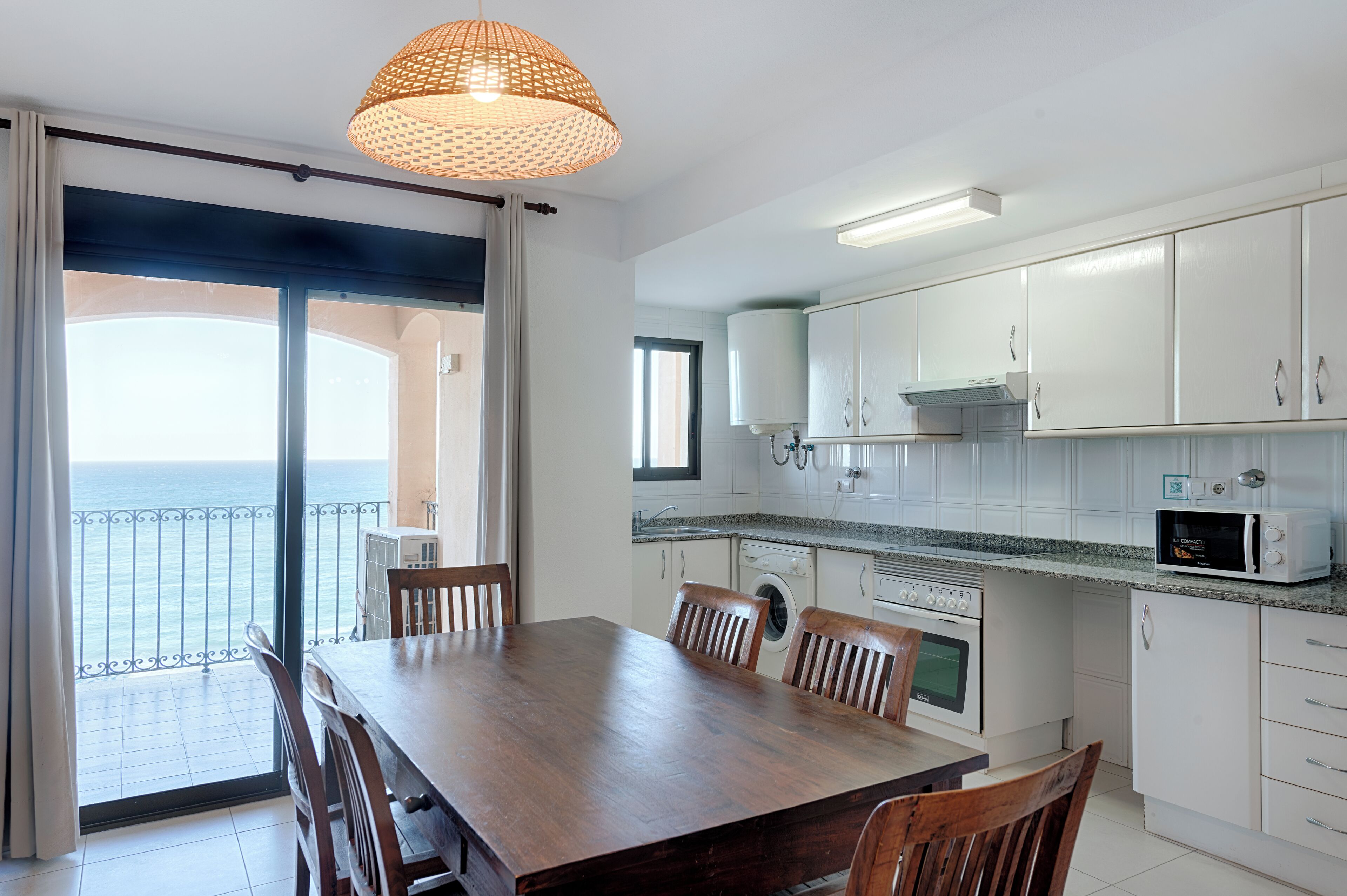 A dining area with a table and chairs, a kitchen with white cabinets, a microwave, and a view of the sea through a sliding door.