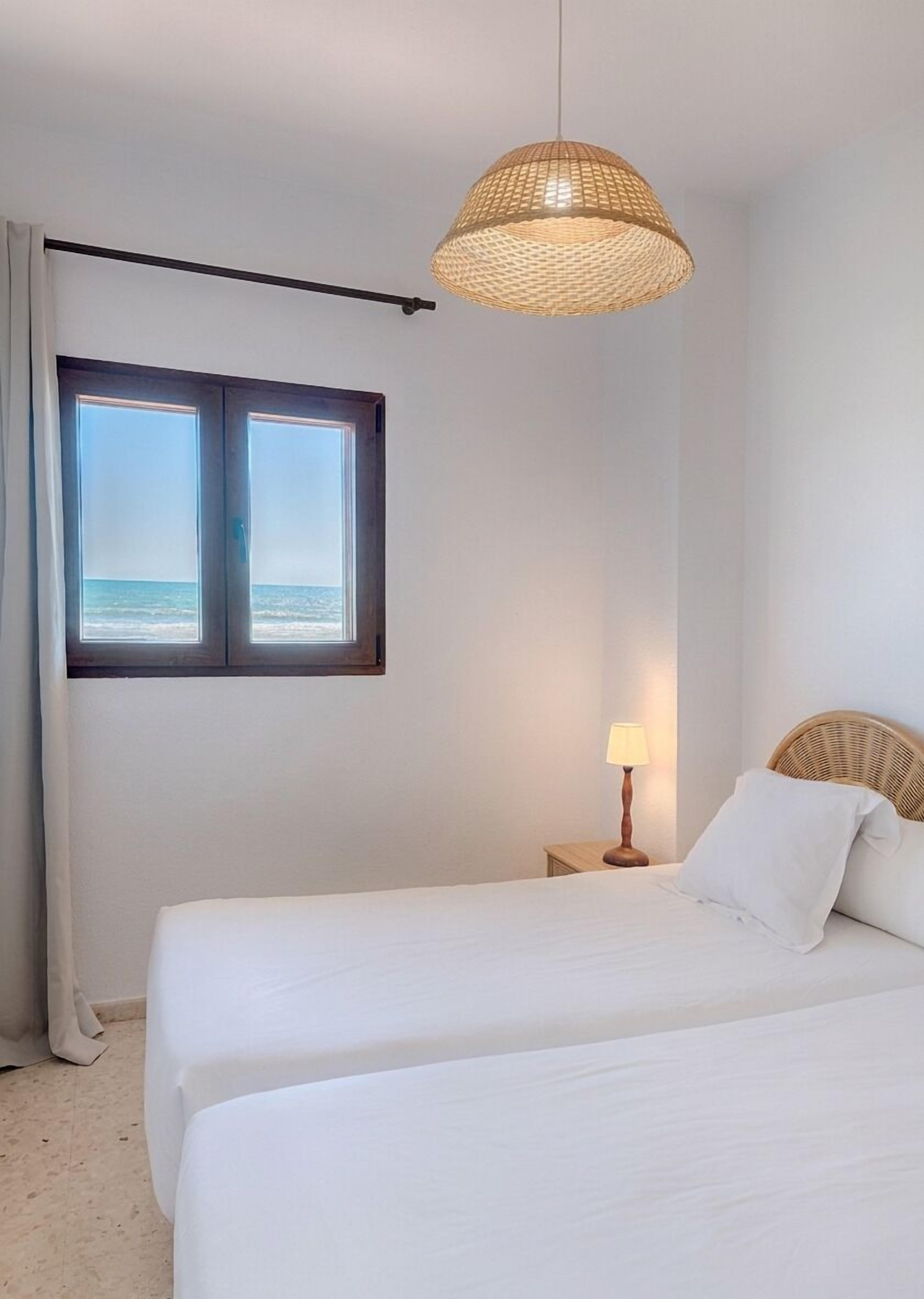 A hotel room with two beds, a window with a view of the sea, a woven ceiling light, and a bedside table with a lamp.