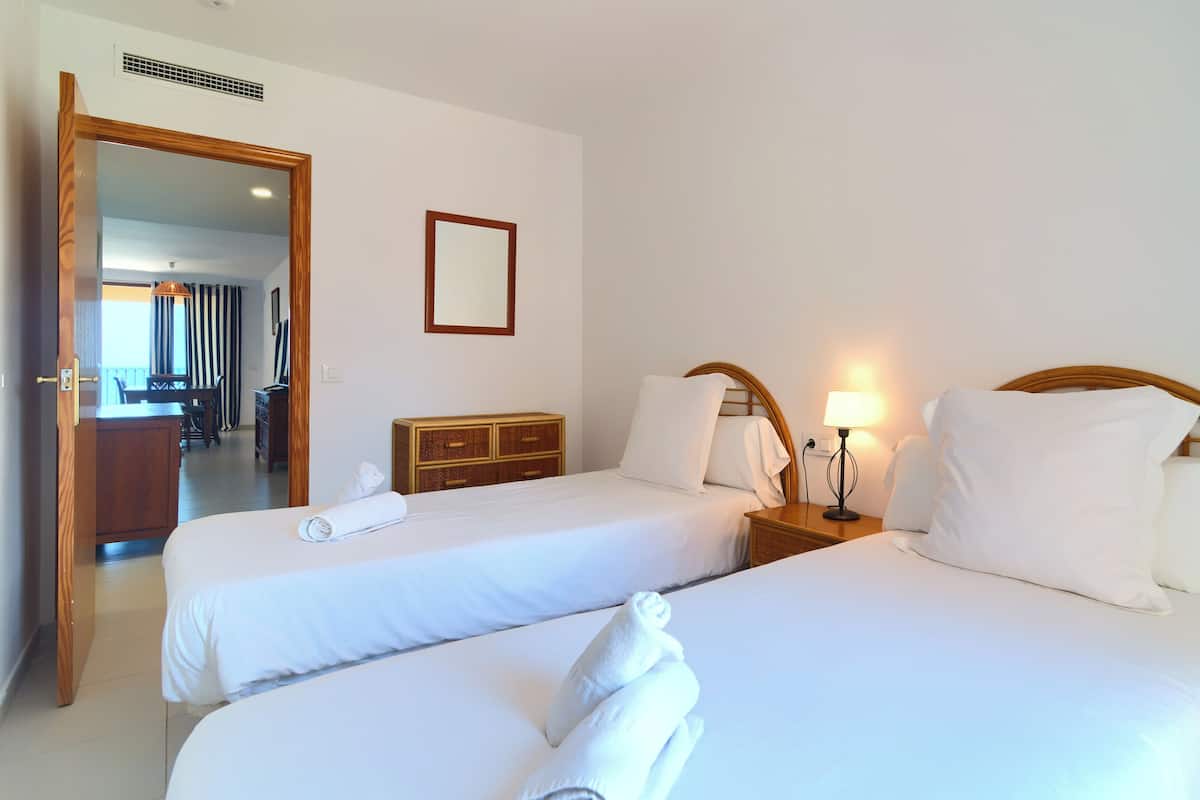 Quarto de hotel com duas camas, cómoda de madeira, espelho e secretária com cadeiras.