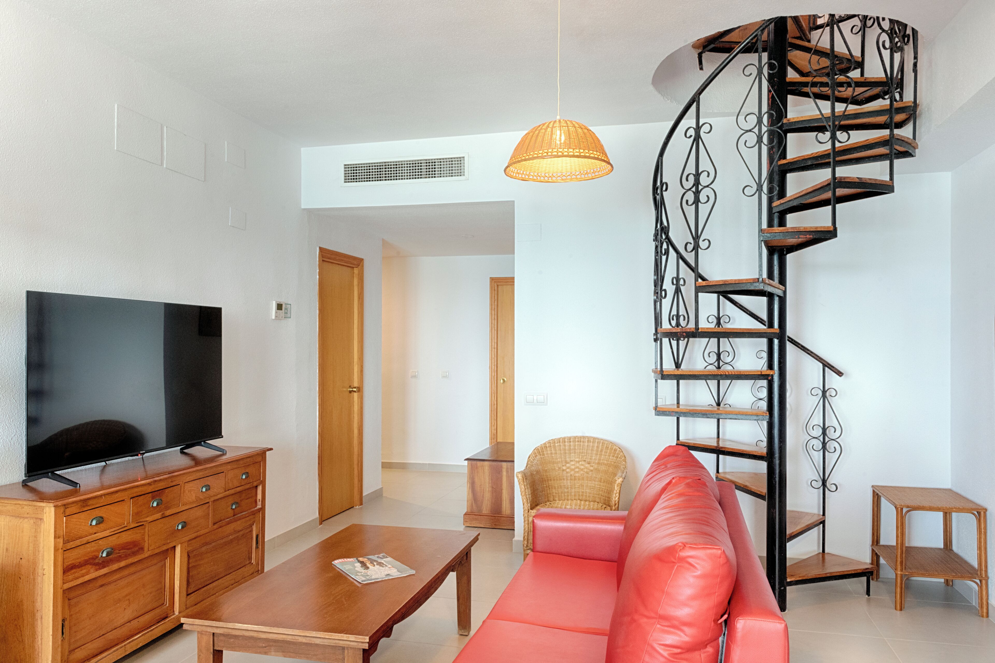Un salon équipé d’un téléviseur, de meubles en bois, d’un canapé rouge, d’un fauteuil en osier, d’une table basse en bois, d’une lampe en osier et d’un escalier en colimaçon.