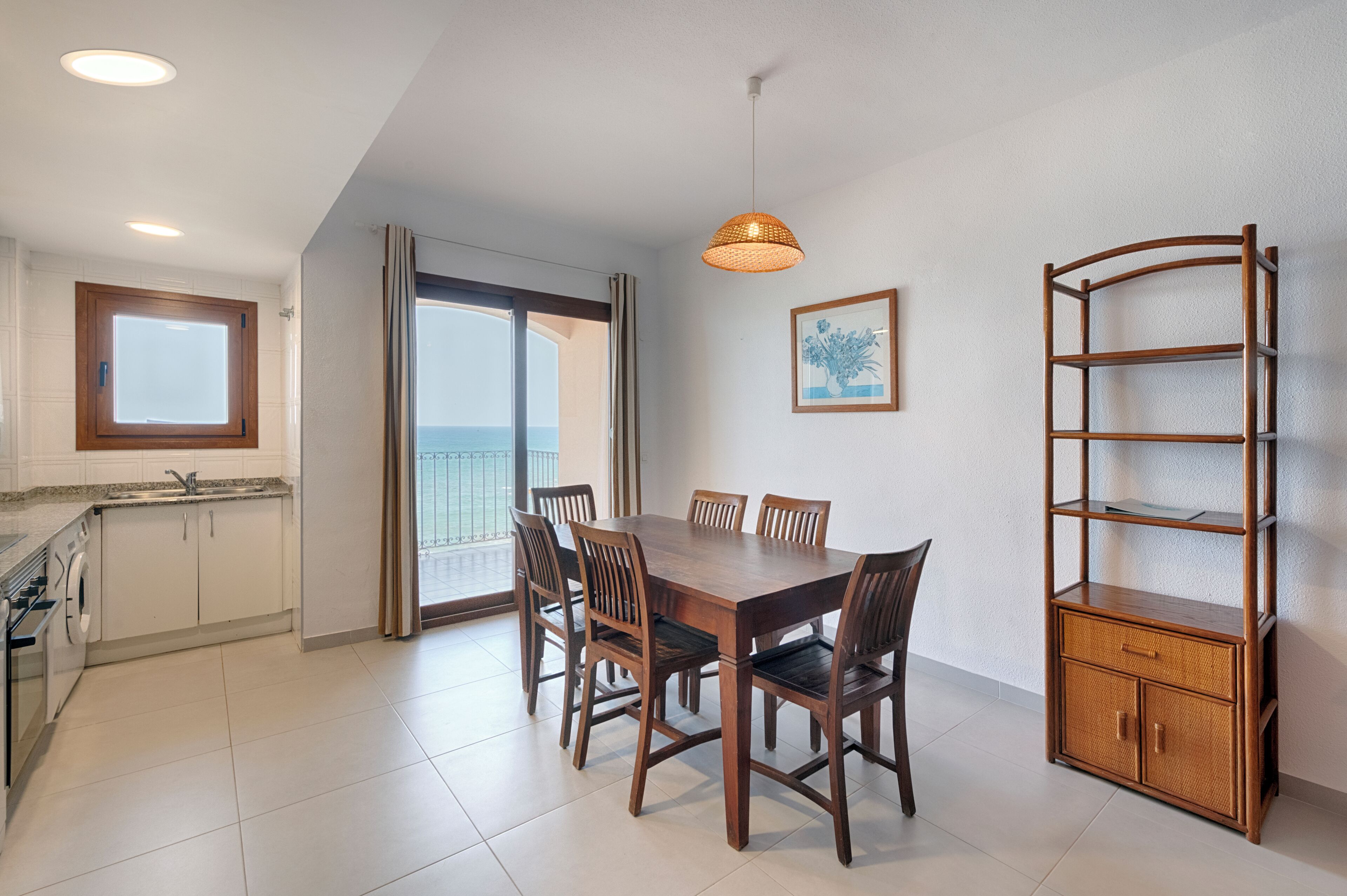 A dining area with a wooden table and chairs, a hanging light fixture, a kitchen with a window, and a view of the outdoors.