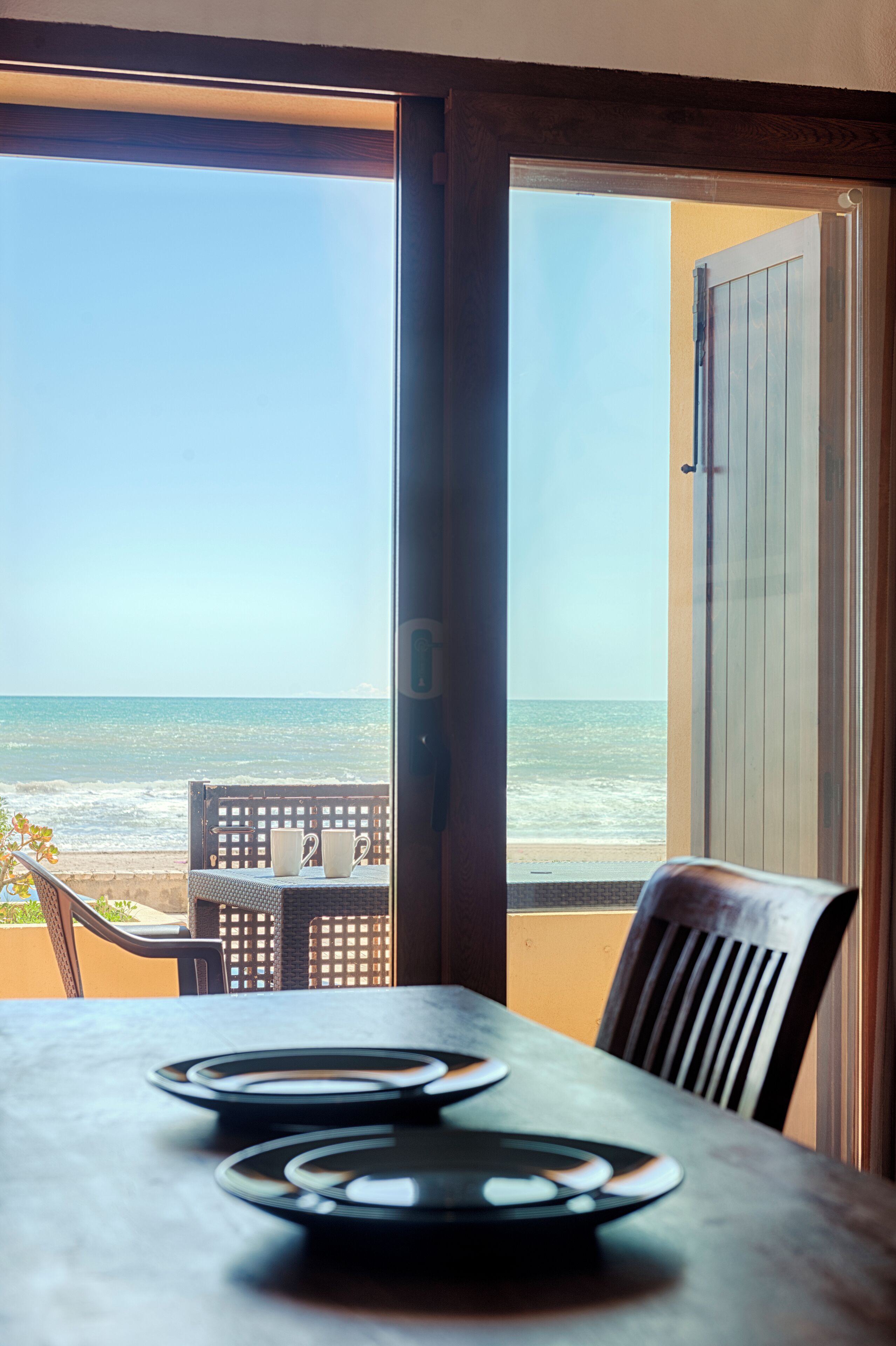 A table with plates and a chair, a view of the beach and ocean through an open door.