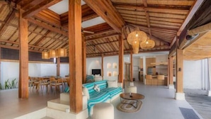 A spacious indoor area with a high ceiling, wooden beams, a sofa set, a dining table, and a kitchen area. - Villa Elora Canggu (Bali)