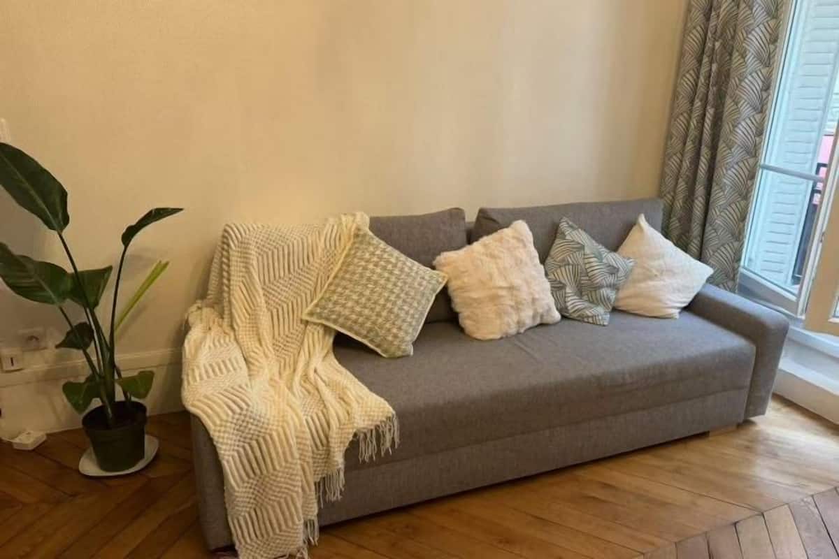 A grey sofa with a white throw blanket and patterned pillows, a potted plant, and a window with curtains.