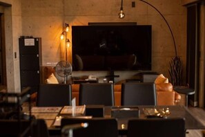 A restaurant with a large TV screen, tables, chairs, and a fan.