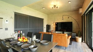 Ein moderner Essbereich mit einem langen Tisch, der für eine Mahlzeit gedeckt ist, ein Küchenbereich mit einem großen Fenster und eine Sitzecke mit einer Couch und einem Fernseher.