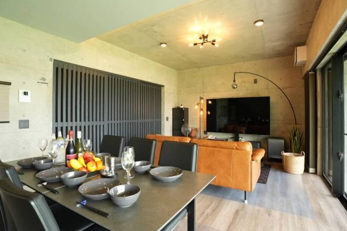 Un espace repas moderne avec une longue table dressée pour un repas, un coin cuisine doté d’une grande fenêtre et un coin salon avec un canapé et un téléviseur.