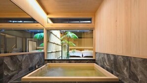 Un espace de spa moderne avec un plafond en bois, un grand bain à remous rectangulaire et une vue sur une pièce de style japonais traditionnel, dotée de tatamis et d’un paravent shoji.