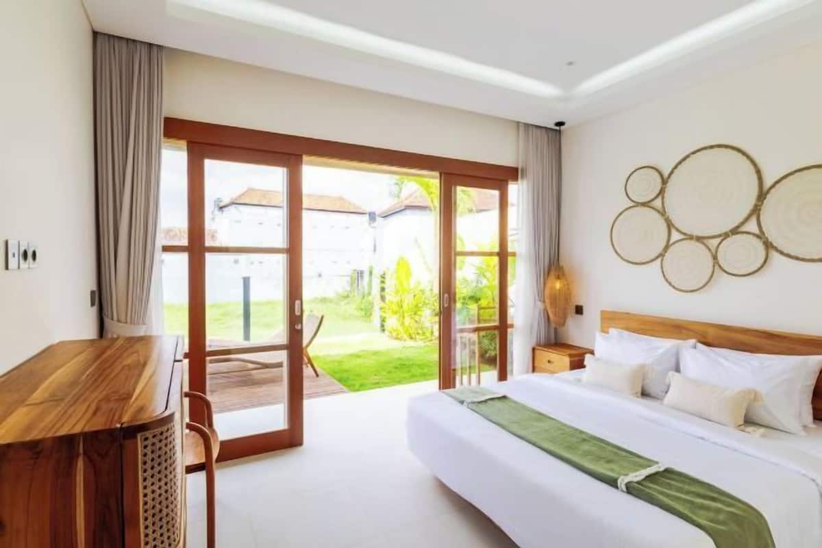 A bedroom with a bed, a desk, a chair, a nightstand, and a view of the outdoors.
