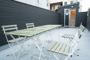 Un espace extérieur aménagé avec une table et des bancs, un mur sombre et l’entrée d’un bâtiment.
